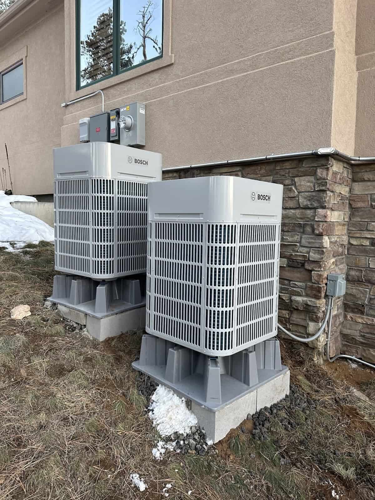 Two Bosch heat pumps on raised bases beside a house after snowfall