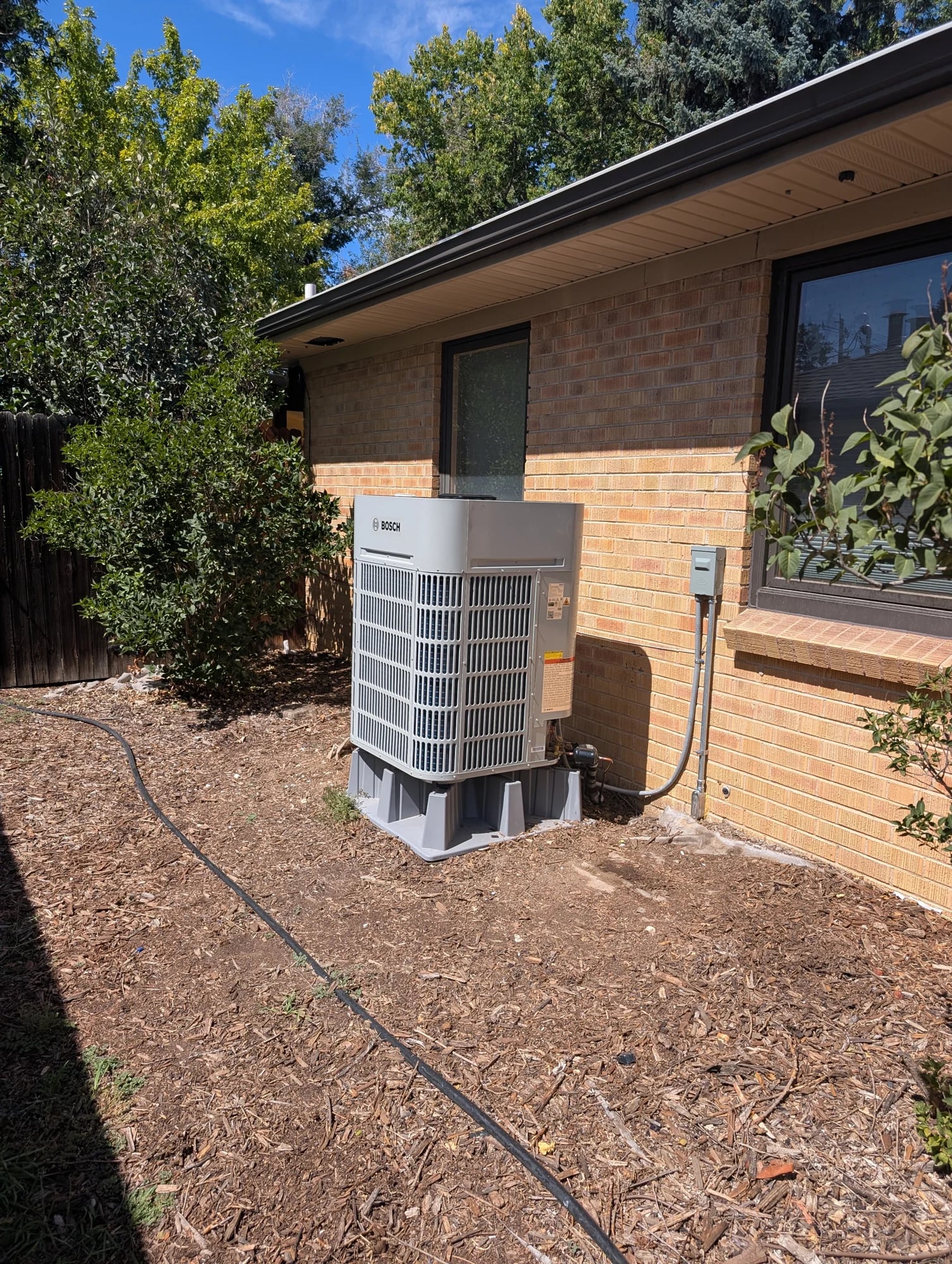 Bosch outdoor heat pump on a raised base beside a tan brick house, with a disconnect box on the wall