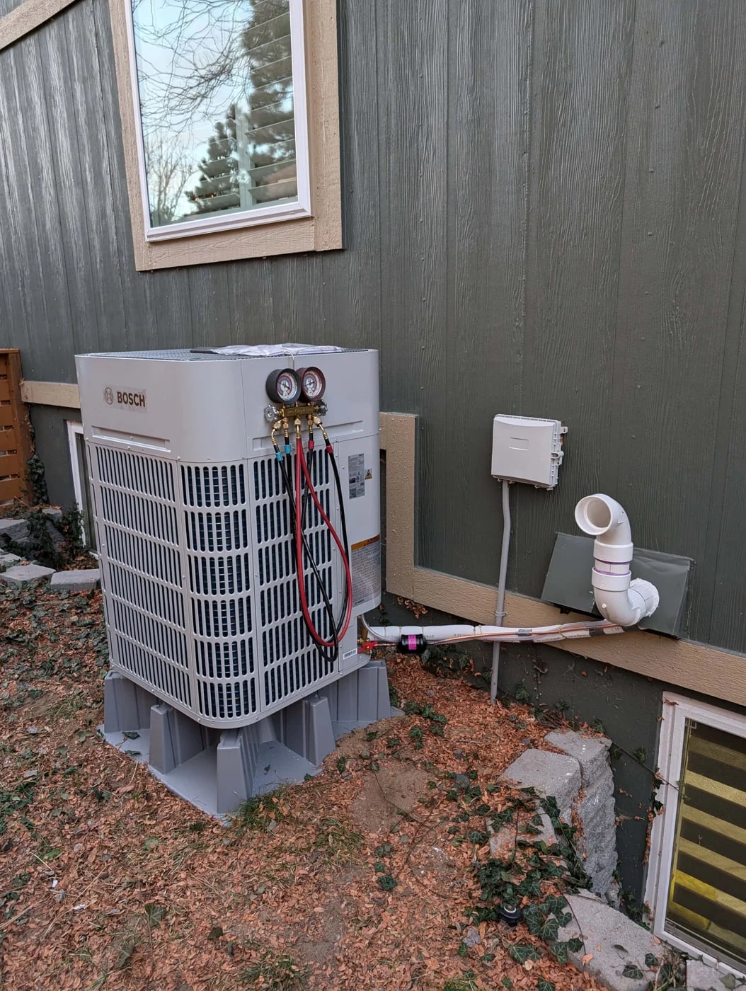 Red and blue refrigerant gauges connected to a Bosch outdoor unit beside green siding
