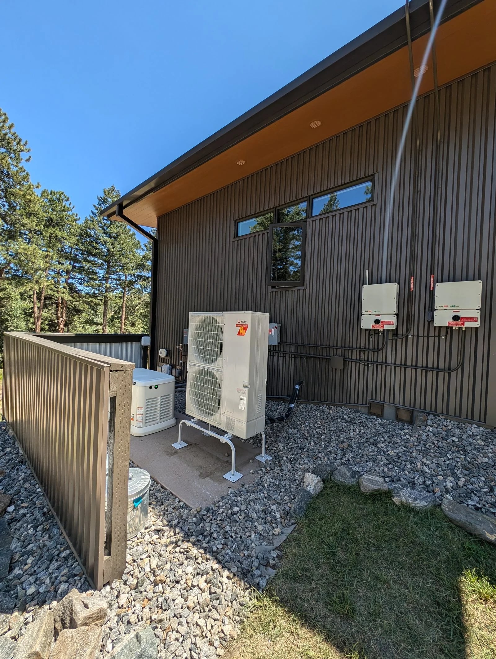 A Mitsubishi heat pump on a stand behind a metal privacy screen at a dark-sided house among pine trees