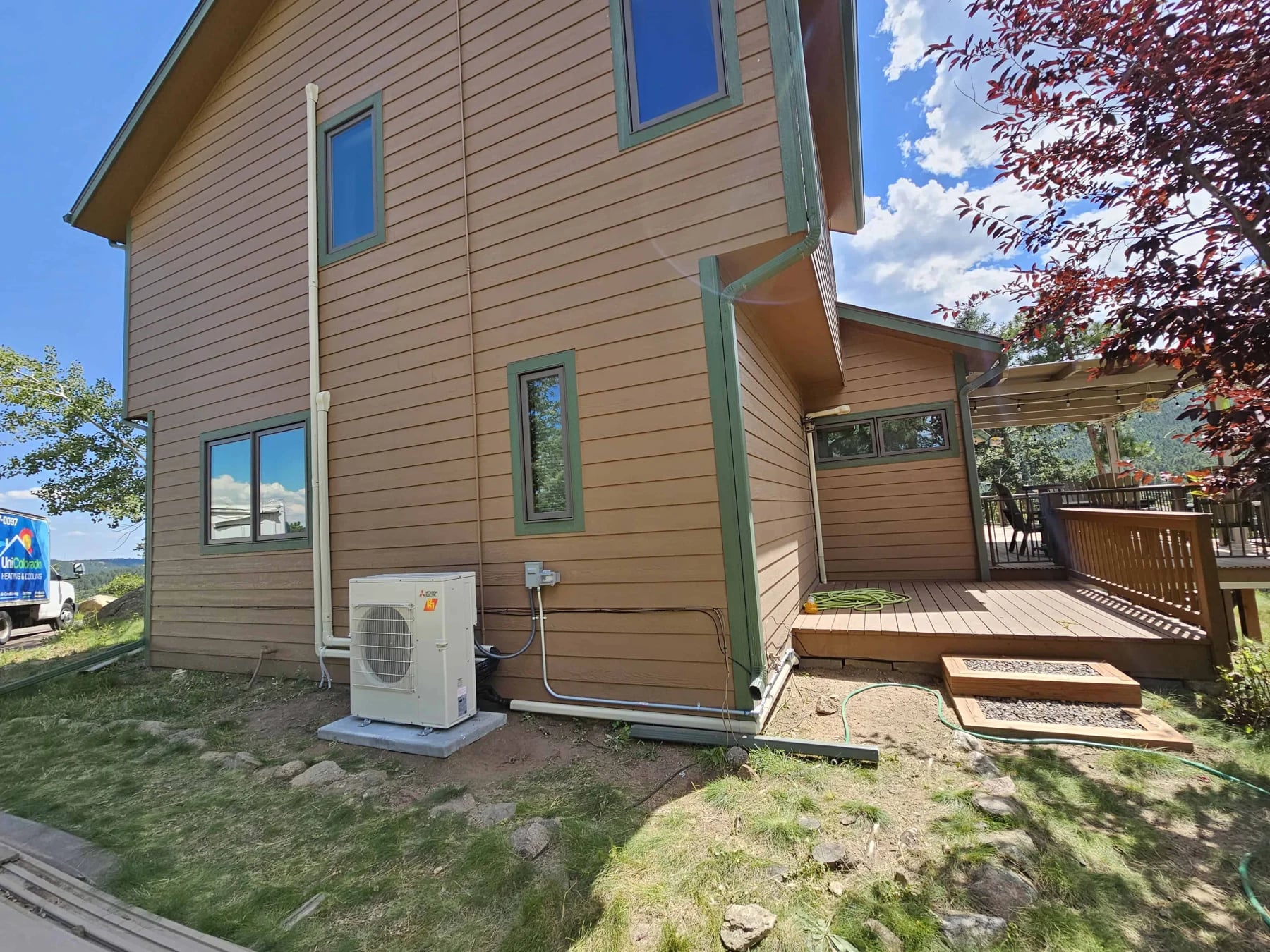 Ductless outdoor unit on a pad beside tan siding, with a wall-mounted disconnect and conduit, near a wooden deck