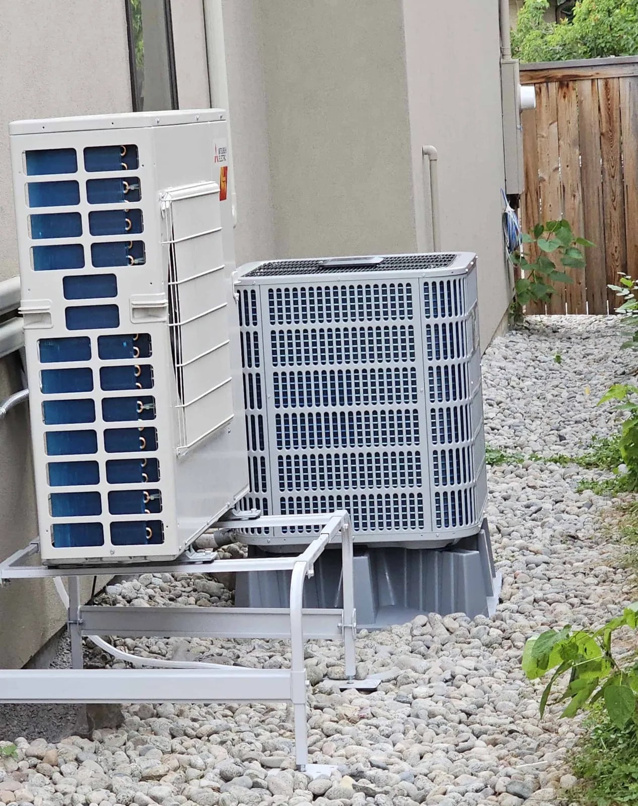A Mitsubishi ductless outdoor unit on a wall stand next to a central heat pump on a riser pad, over river rock beside stucco