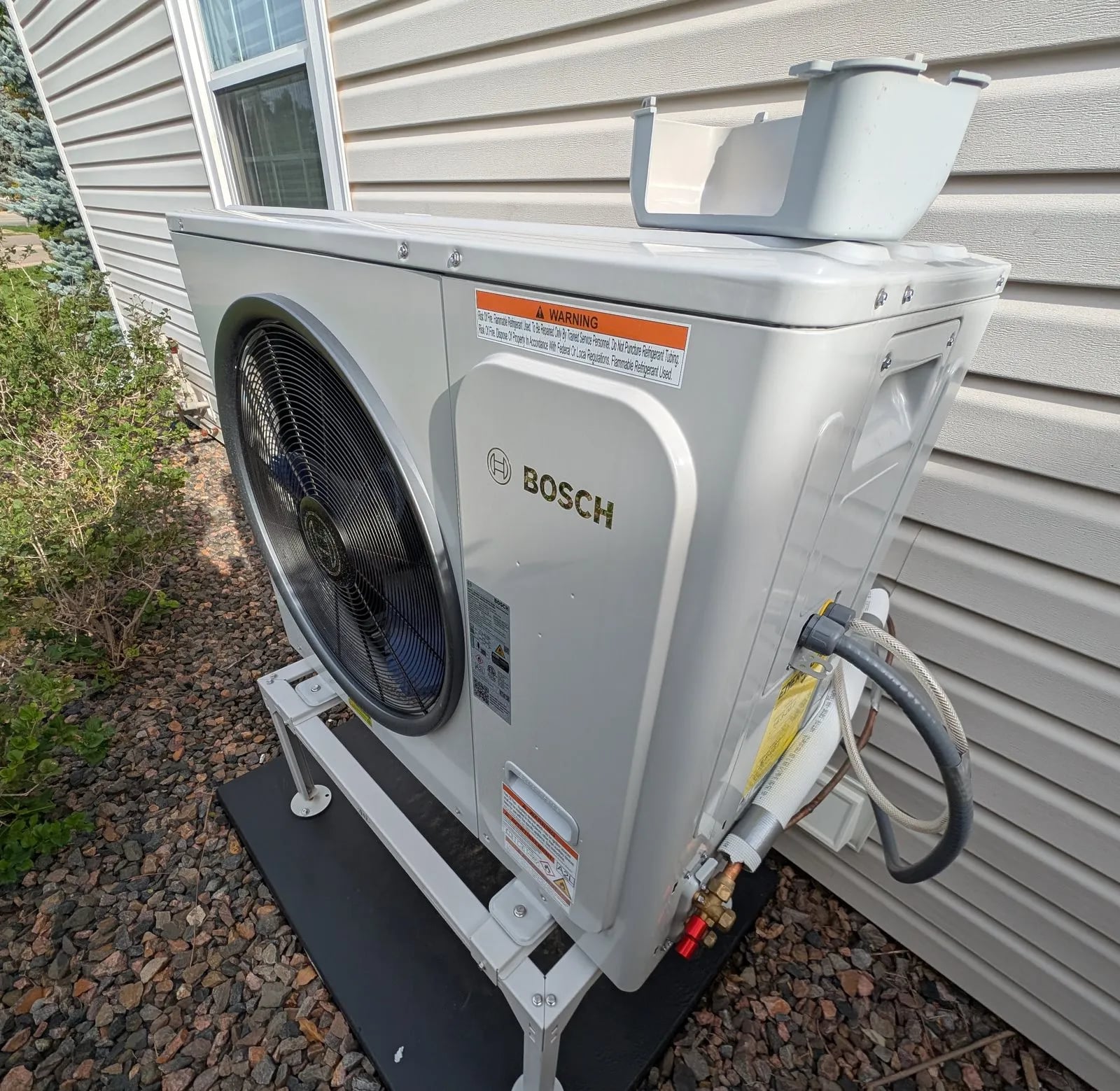 A white Bosch IDS heat pump outdoor unit on a stand over mulch beside beige siding