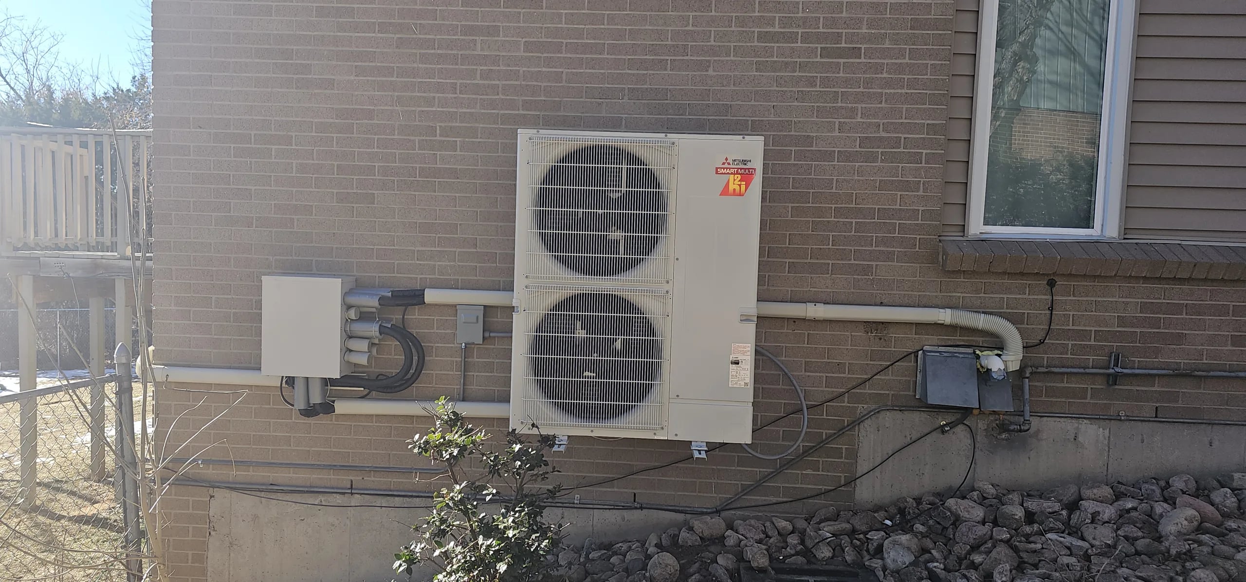 A two-fan Mitsubishi outdoor unit on a wall bracket on a brick wall, with a line set cover and a disconnect