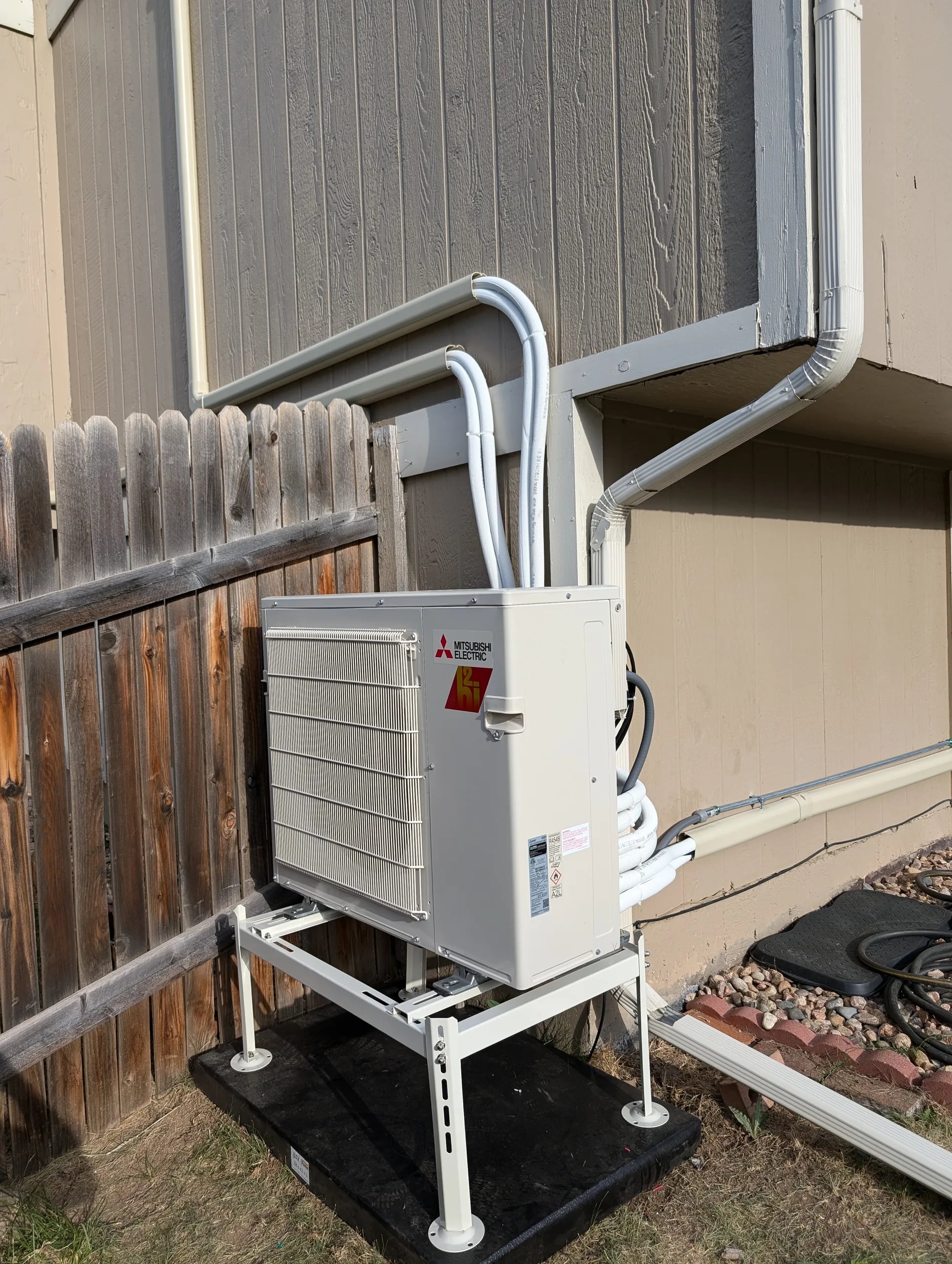 A Mitsubishi outdoor unit on a stand over a rubber mat beside a wood fence, with line set covers on the wall above