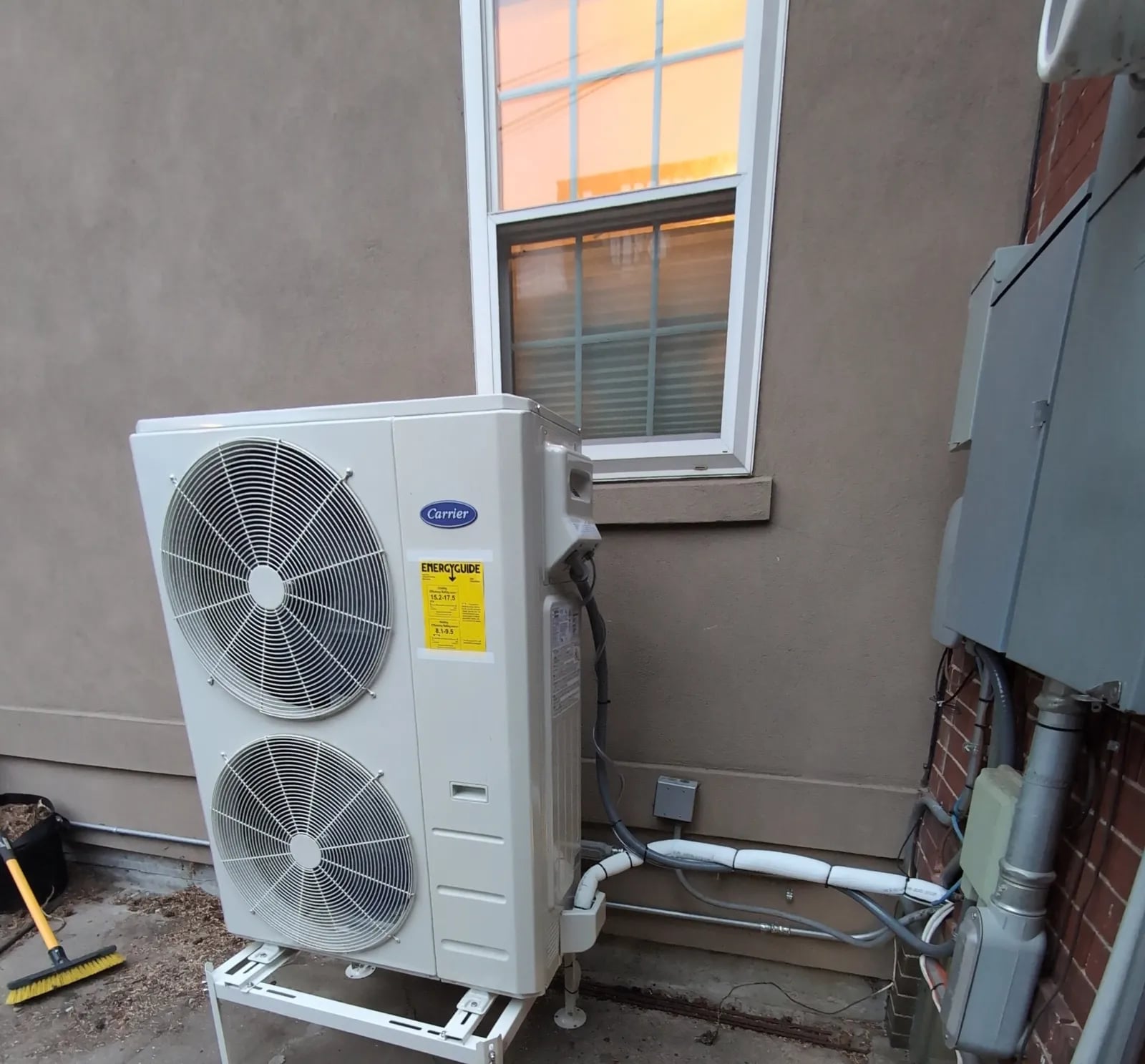 A Carrier two-fan heat pump outdoor unit on a stand beside a stucco wall below a window