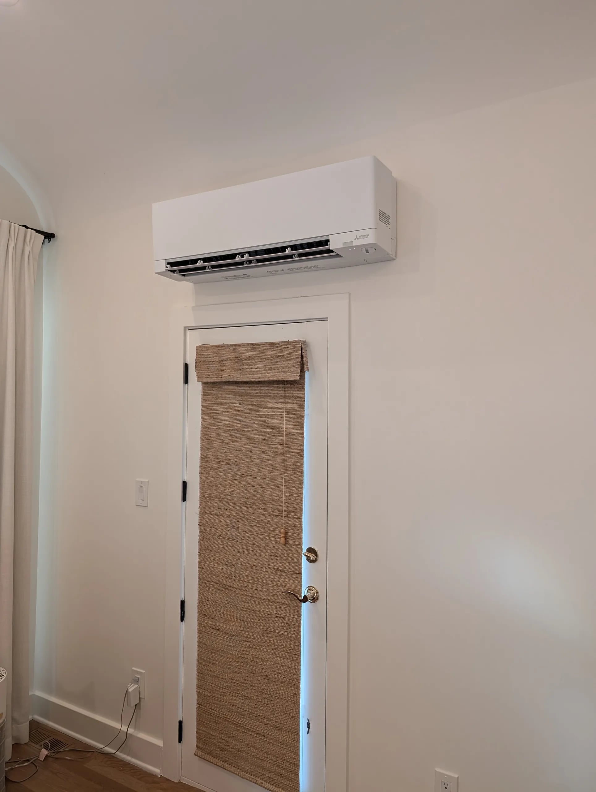 A white Mitsubishi wall-mounted head above a door with a woven shade in a white room