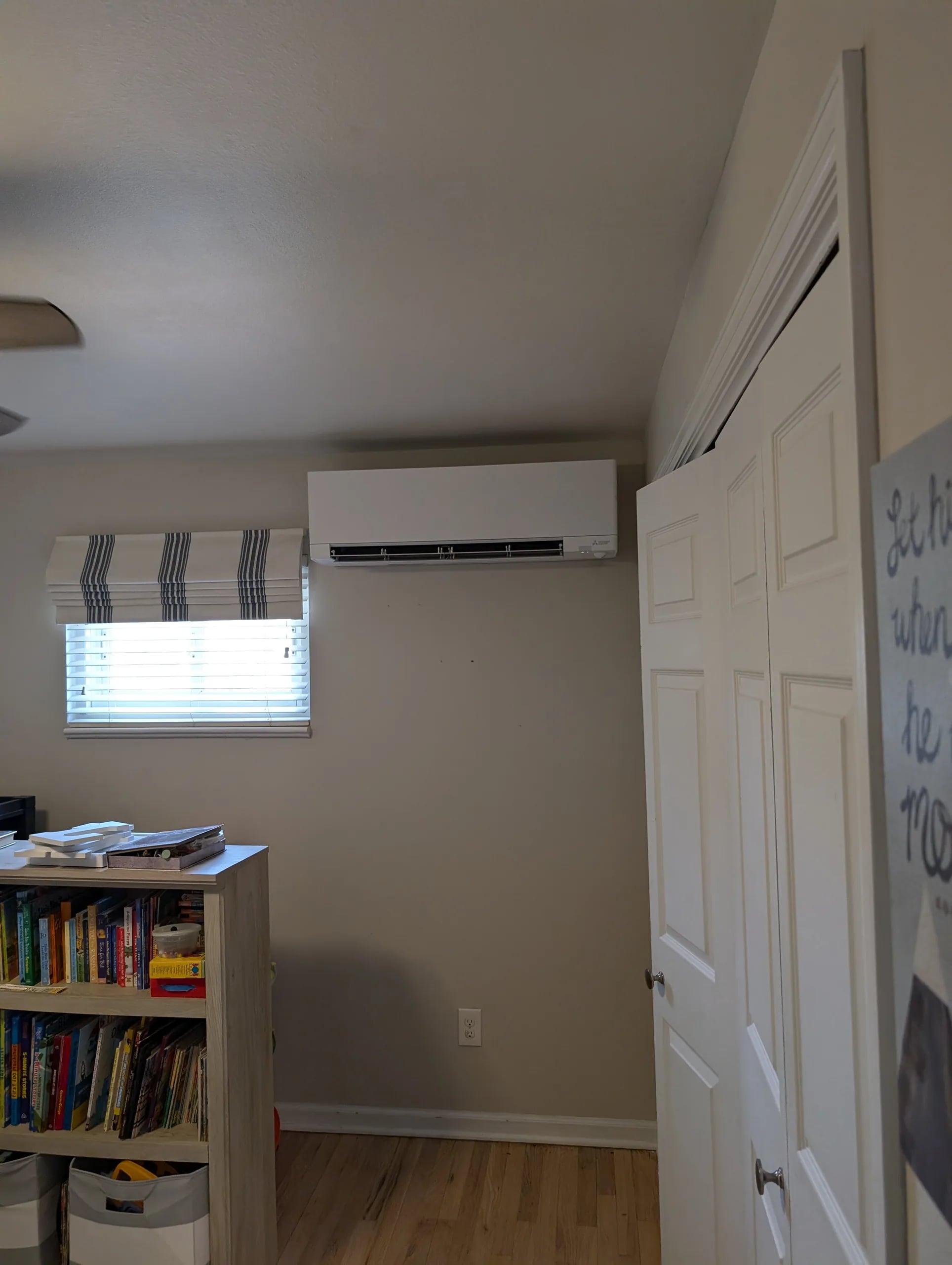 A white ductless wall head in a child's room above a bookshelf and a striped window shade