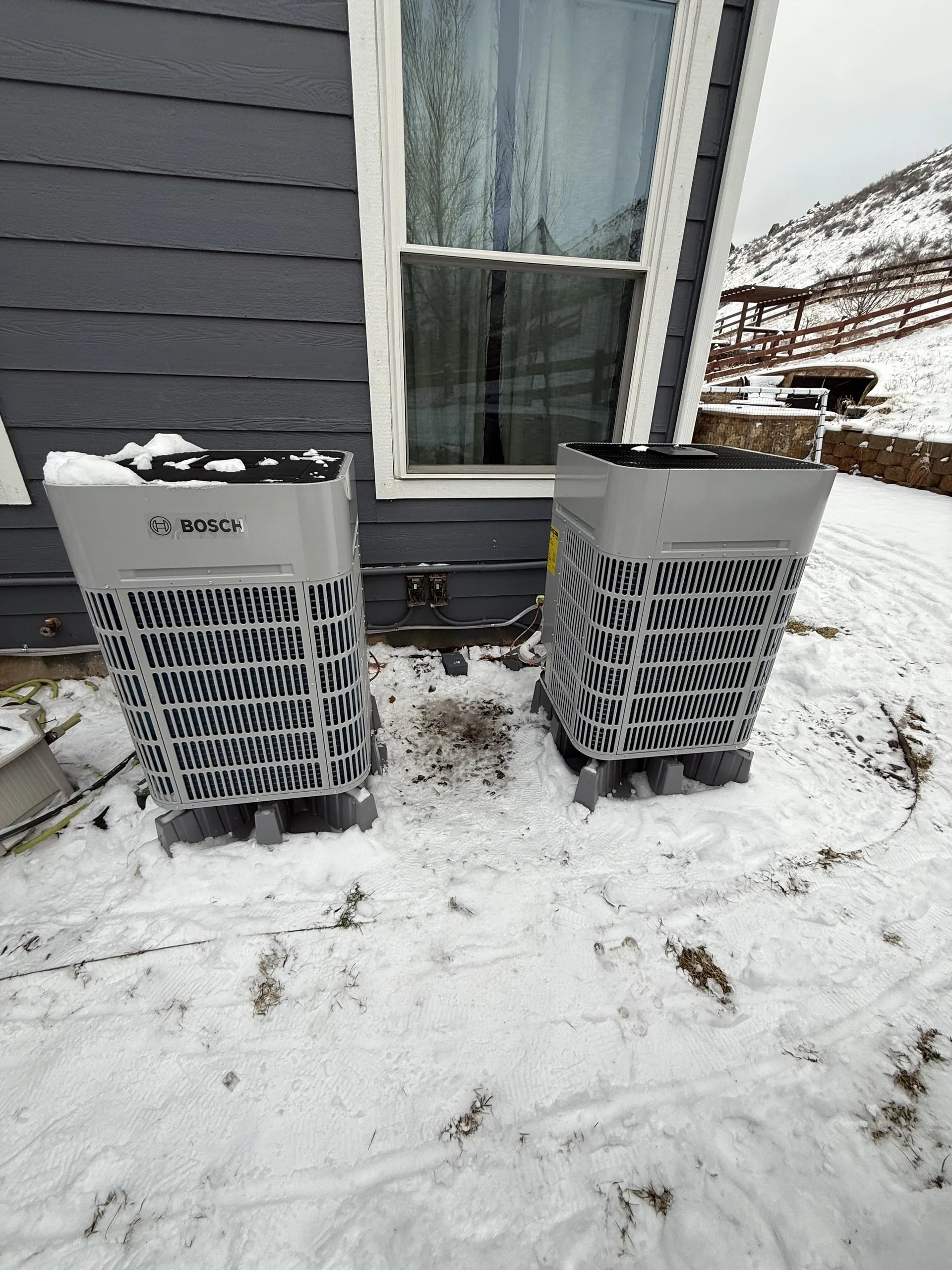 Two Bosch outdoor units on risers beside dark siding, with snow on the ground