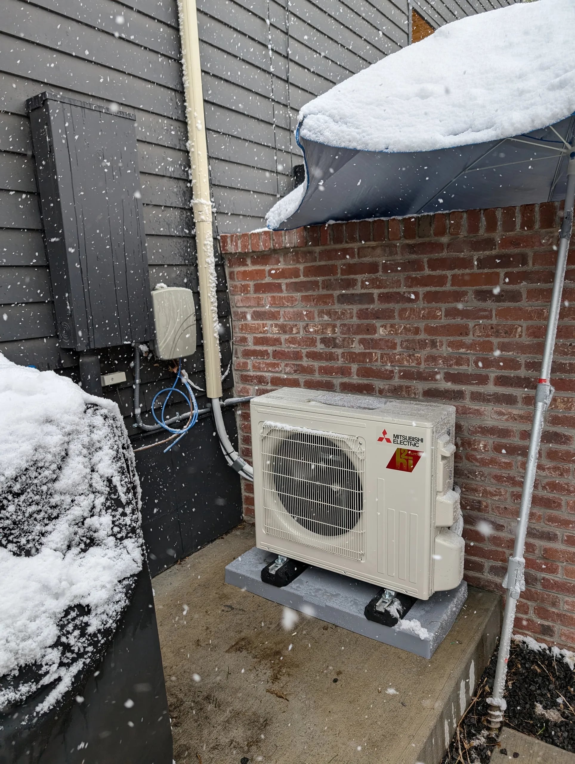 A compact Mitsubishi outdoor unit on a pad beside a red brick wall, with snow on the shrubs and a covered line set above