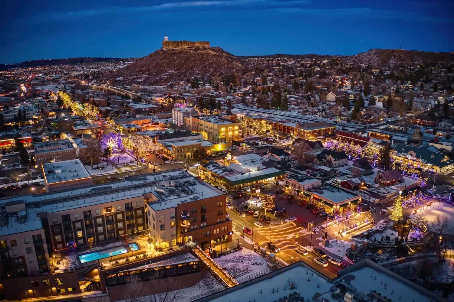 Aerial dusk view of a snowy Front Range town center lit up below a flat-topped butte