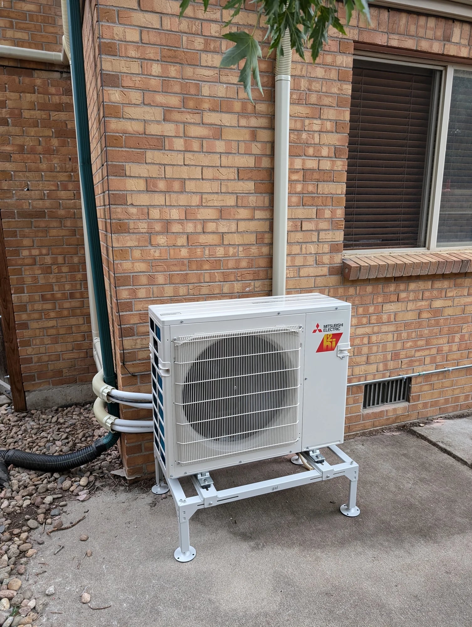 A Mitsubishi outdoor unit on a galvanized stand on concrete beside a brick wall